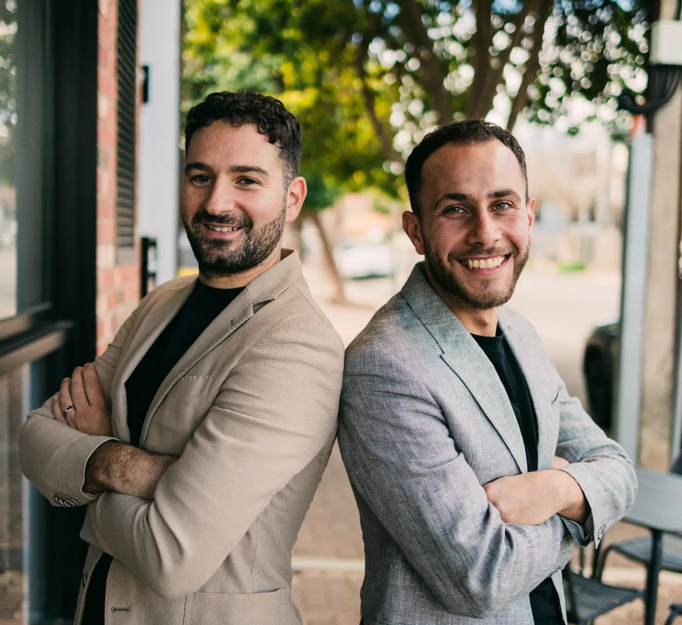 Two men in suits standing back-to-back outdoors with trees and a building in the background.